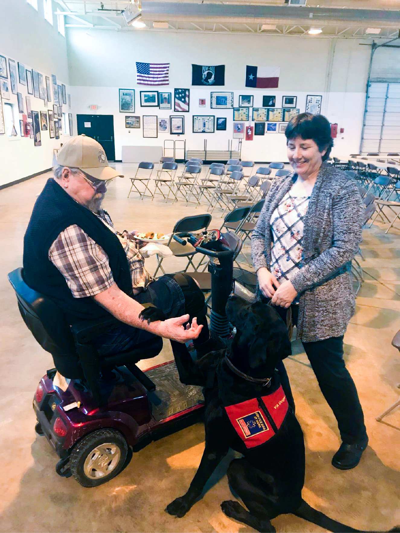 A man sitting on a mobility scooter interacts with a black service dog wearing a red vest indoors, while a woman stands nearby smiling. Behind them are several flags and framed photos on the walls. The room contains rows of empty chairs.