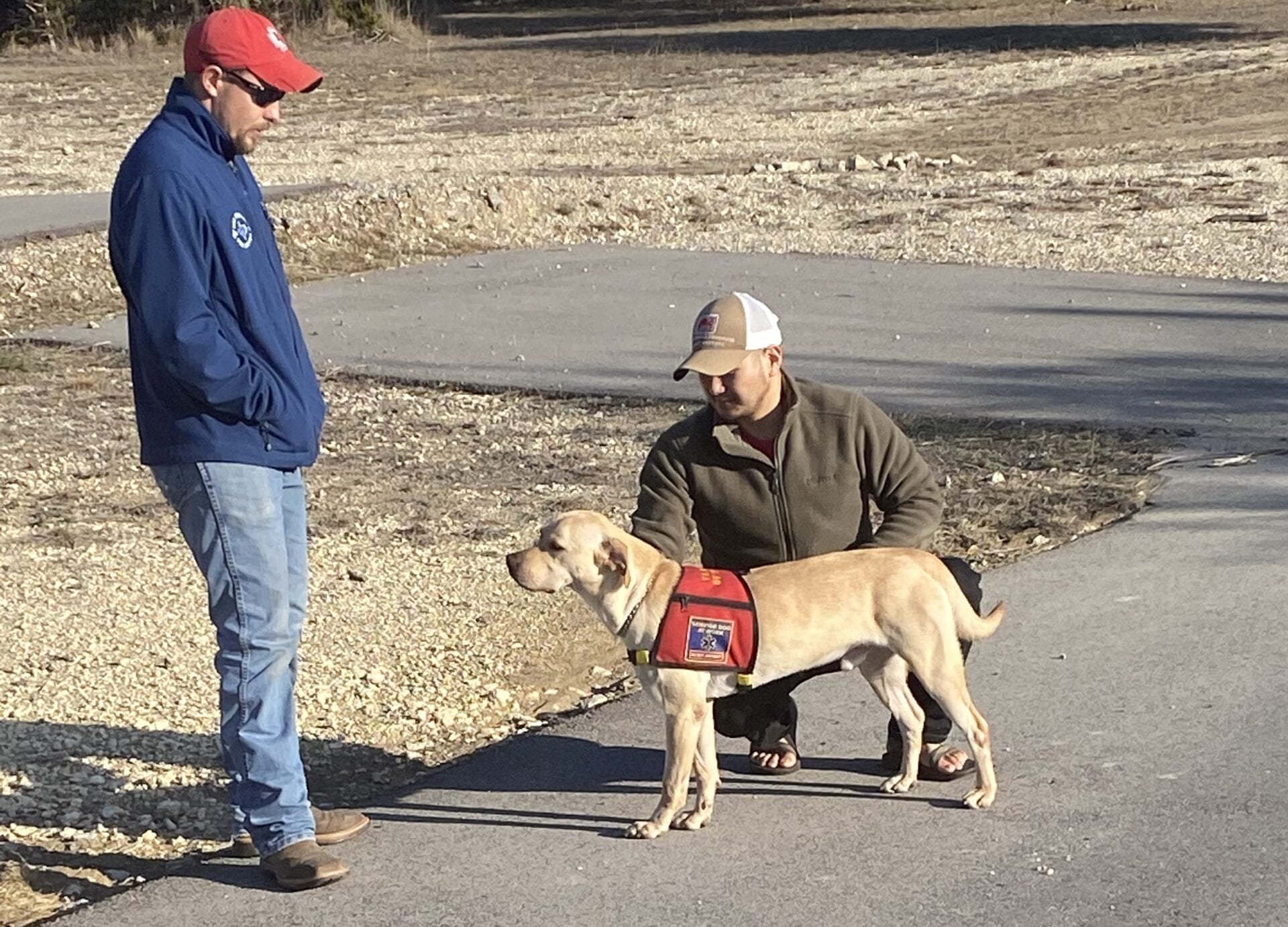 Two men interacting with a Labrador wearing a red service vest on a paved pathway outdoors.