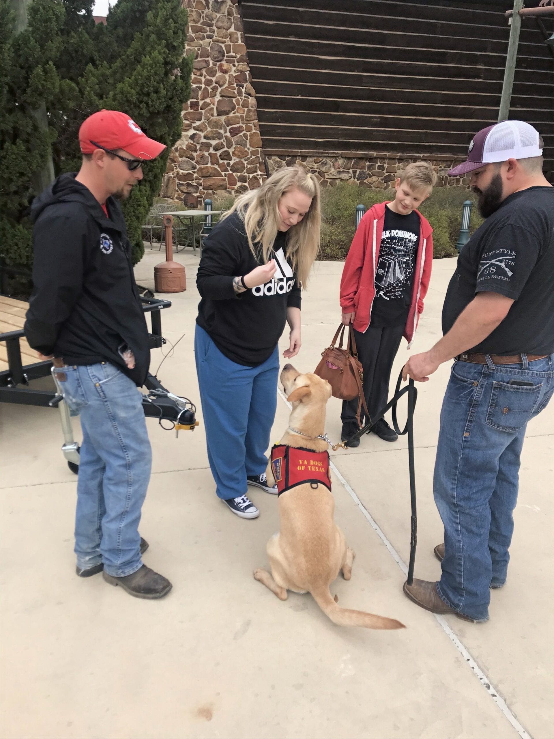 Group of people interacting with a service dog wearing a 'VA Dogs of Texas' vest outdoors.