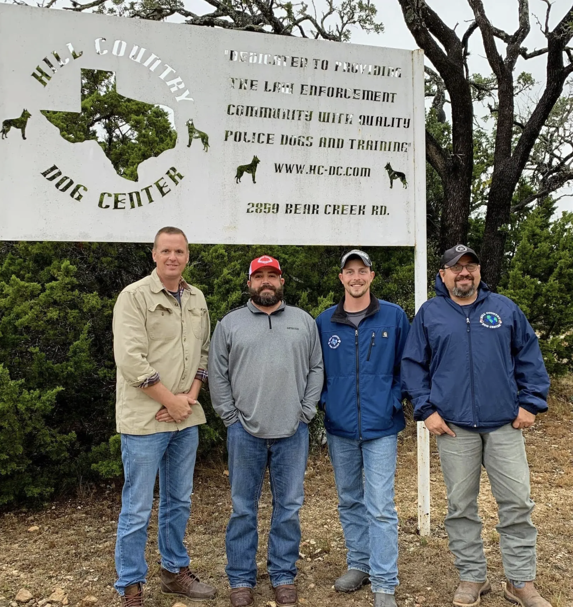 Four men standing in front of a sign for Hill Country Dog Center, which provides quality police dogs and training, located at 2859 Bear Creek Rd.