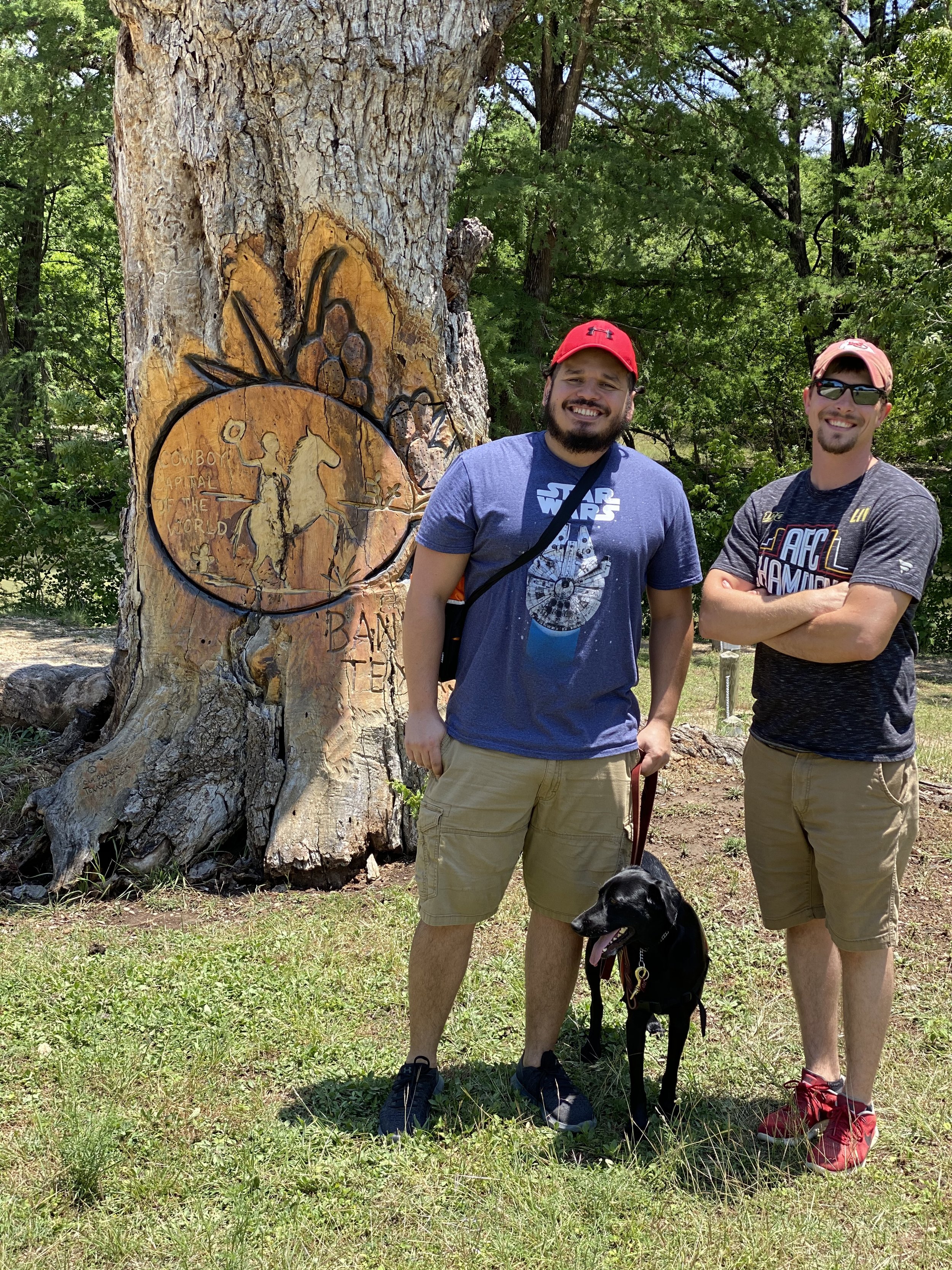 Two men standing in front of a large tree with carved artwork, accompanied by a black dog, in a grassy outdoor setting.